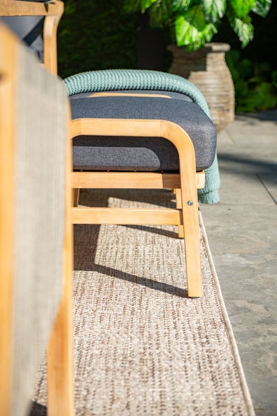 Nahaufnahme eines Gartenstuhls aus Holz mit grauem Polster und einer Decke auf einem gewebten Outdoor-Teppich auf einer Terrasse.