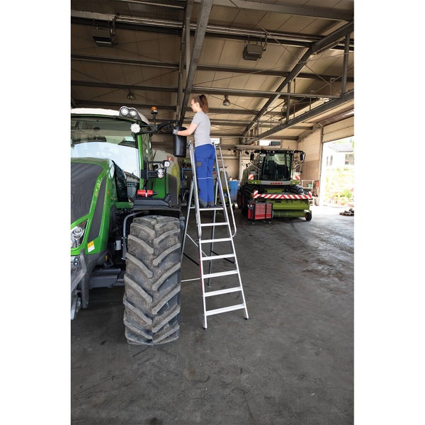 Person auf einer Aluminium Stufenstehleiter bei der Wartung eines Traktors in einer geräumigen Werkstatt.