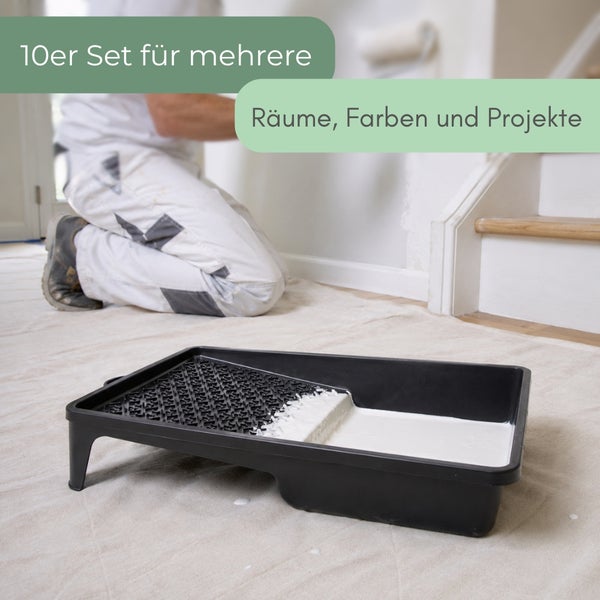 Schwarze Farbwanne mit weißer Farbe auf Malervlies, im Hintergrund ein kniender Maler in einem hellen Raum.