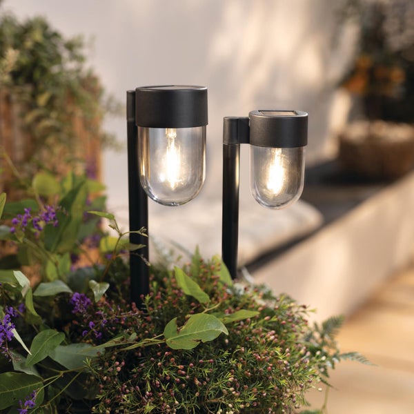 Zwei schwarze Solar-LED-Erdspießleuchten mit warmweißem Licht in einem Pflanzgefäß mit grünen Blättern.