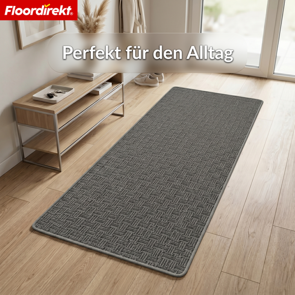 Grauer Läufer mit Strukturmuster in einem hellen Flur auf Holzboden neben einem Konsolentisch. Floordirekt Logo.
