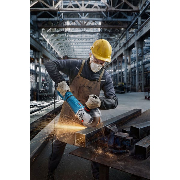 Ein Arbeiter mit Schutzhelm und Schutzmaske schleift Metall mit einem Winkelschleifer in einer Werkshalle.