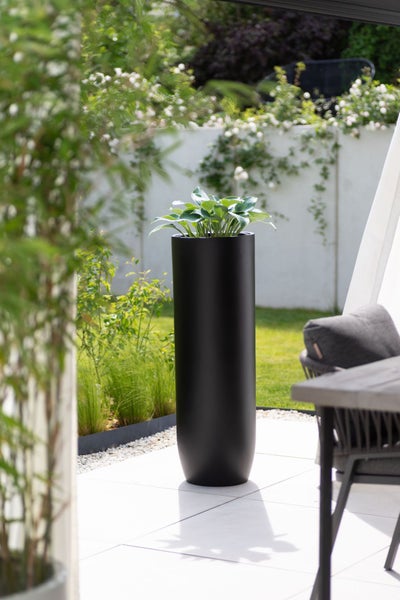 Hoher, schwarzer Pflanzkübel mit grüner Pflanze auf einer modernen, hellen Terrasse in einem gepflegten Garten.