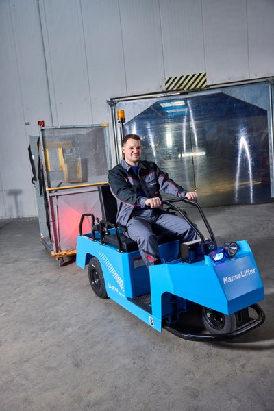 Blauer HanseLifter Elektro-Schlepper mit Lithium-Ionen-Batterie, bedient von einem Mann in einem hellen Lager beim Ziehen eines Gitterwagens.