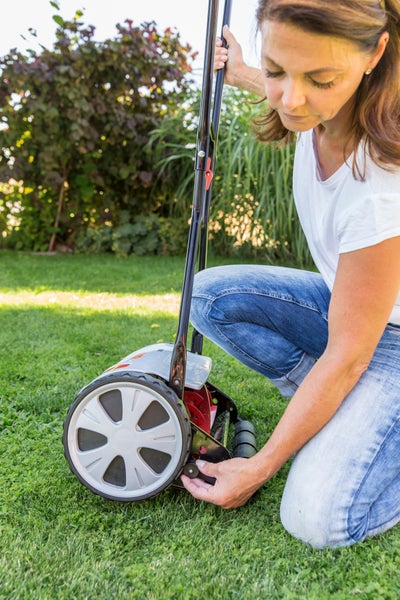 Eine Frau kniet im Garten und stellt die Schnitthöhe eines Handspindelrasenmähers ein.