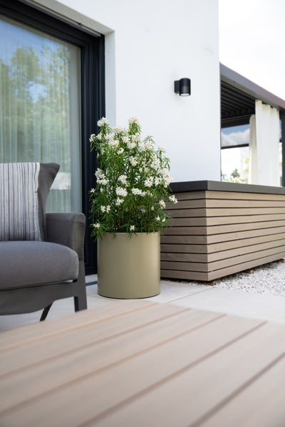 Moderner runder Pflanzkübel in Olivgrün auf einer hellen Terrasse neben einem grauen Sessel und einer Holzbank.