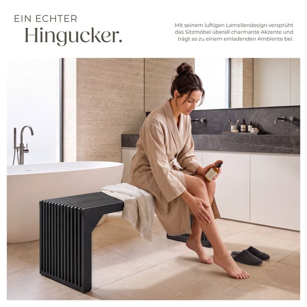 Frau im Bademantel sitzt auf einer modernen Sitzbank mit Lamellendesign in einem hellen Badezimmer mit Badewanne und Waschbecken.