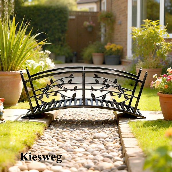 Dekorative Gartenbrücke aus Metall mit Blattmotiv, aufgestellt über einem Kiesweg in einer hellen Gartenanlage mit Rasen und Pflanzen.