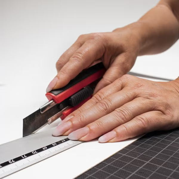 Eine Hand schneidet mit einem Cuttermesser und einem Metalllineal auf einer Schneidematte ein Stück Papier.