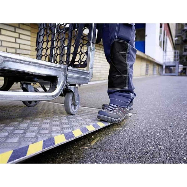 Person in Arbeitshose und Sicherheitsschuhen steht neben einem Transportwagen auf einer Rampe mit Warnmarkierung.