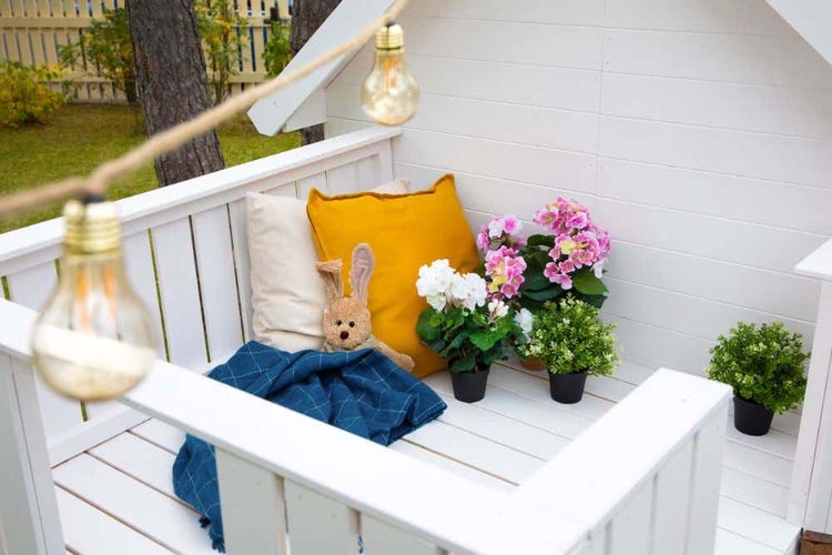 Weißes Garten-Spielhaus aus Holz mit Balkon, dekoriert mit Kissen, Kuscheltier, Decke, Topfpflanzen und Lichterkette.