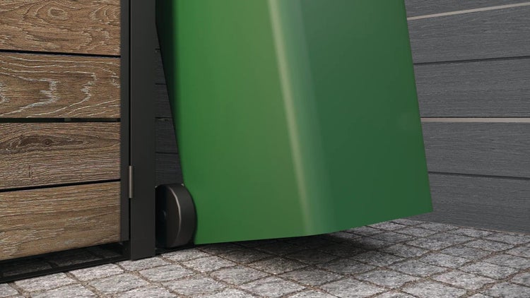 Grüne Mülltonne mit Rollen neben einem Holzzaun auf Kopfsteinpflaster.