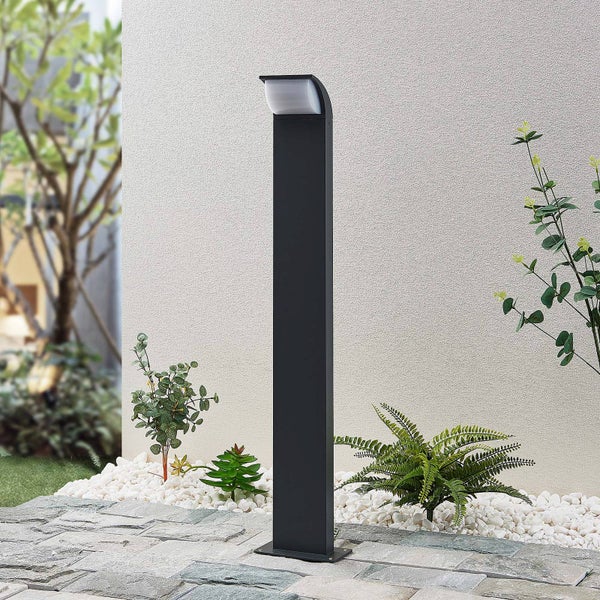 Moderne schwarze Außenleuchte in Säulenform auf einer Steinterrasse vor einer hellen Hauswand mit kleinen Gartenpflanzen.