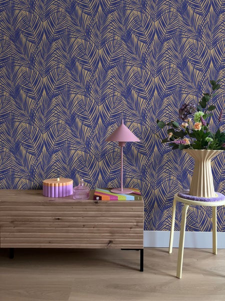 Moderne Innenszene mit Vliestapete im Palmblattmuster. Ein hölzernes Sideboard und ein Hocker mit Blumenvase stehen vor der gemusterten Wand.