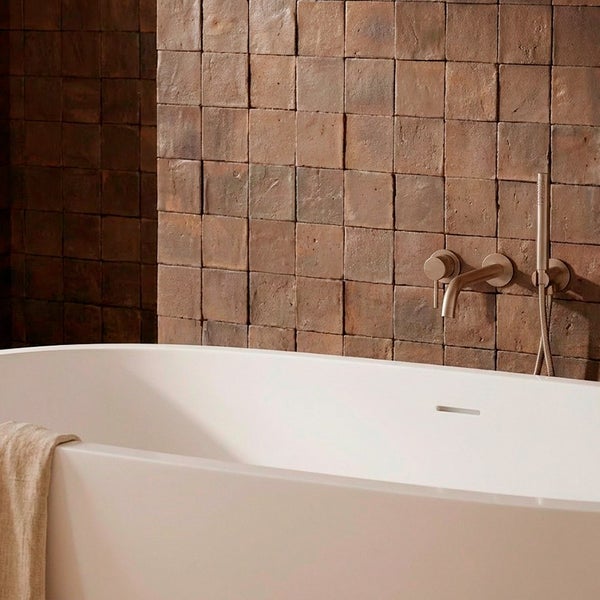 Freistehende Badewanne vor einer Wand mit rustikalen quadratischen Wandfliesen und moderner Unterputz Badarmatur in einem Badezimmer.