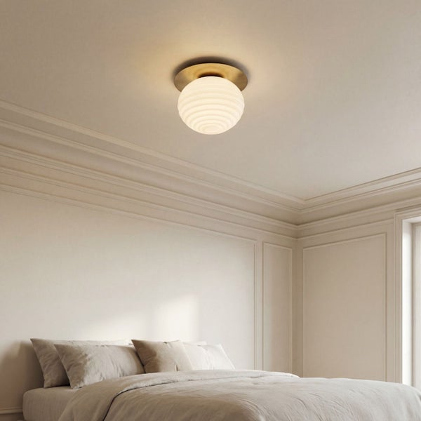 Deckenleuchte mit geriffeltem Glasschirm und Messinghalterung in einem hellen Schlafzimmer mit Wandleisten und Bett.