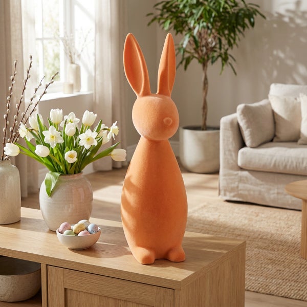 Osterhasen Deko Figur aus beflocktem Material auf einem Holztisch in einem hellen Wohnzimmer mit Blumenvase und Eiern.