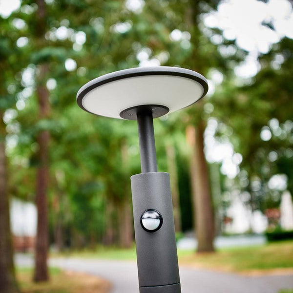 Moderne Außenleuchte mit flachem Kopf und integriertem Bewegungsmelder in einem hellen Garten mit Bäumen und Gehwegen.