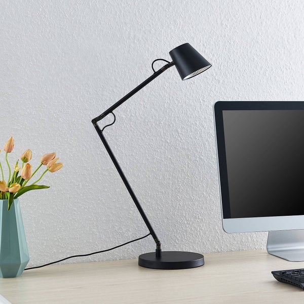 Schwarze Schreibtischlampe aus Metall mit verstellbarem Gelenkarm und rundem Standfuß auf einem Schreibtisch neben einem Monitor.