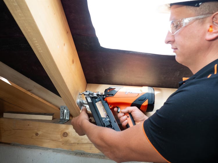 Handwerker befestigt mit einem Akku Nagelgerät einen Metallverbinder an einer Holzkonstruktion auf einer Baustelle.