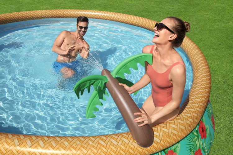 Aufblasbarer Pool mit Rand in Rattan-Optik und aufblasbarer Palme mit Wassersprühfunktion, zwei Personen spielen im Wasser auf einer Rasenfläche.