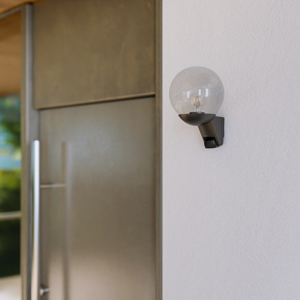 Außenwandleuchte mit kugelförmigem Glasschirm und integriertem Bewegungsmelder an einer Hauswand neben einer Eingangstür.