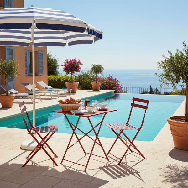 Roter Klapptisch und zwei rote Klappstühle aus Metall auf einer hellen Terrasse neben einem Pool mit blau-weiß gestreiftem Sonnenschirm.