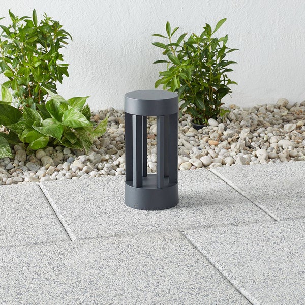 Moderne Gartenleuchte aus anthrazitfarbenem Metall auf hellgrauen Steinplatten in einem Garten mit Kies und Pflanzen.