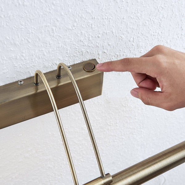 Bilderleuchte aus antikem Messing mit integriertem Schalter am Gehäuse, der von einem Finger bedient wird.