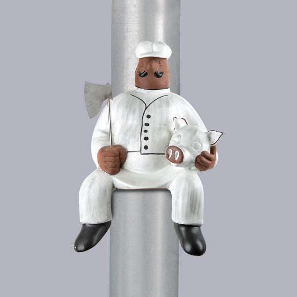 Fallrohrfigur aus Keramik in Form eines Metzgers mit Beil und Schweinekopf, sitzend auf einem Metallrohr.