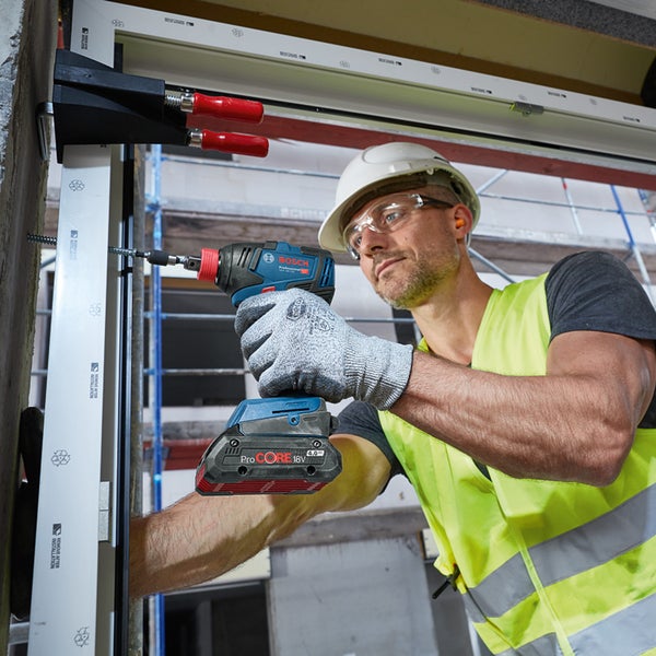 Ein Arbeiter mit Schutzhelm und Warnweste verwendet einen Akku-Schlagschrauber von Bosch, um eine Schraube in einen Fensterrahmen zu drehen.