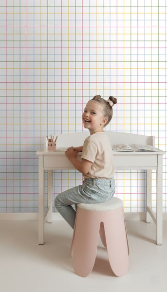 Kinderzimmer mit bunter Gittertapete, weißem Kinderschreibtisch und einem Kind auf einem Hocker.
