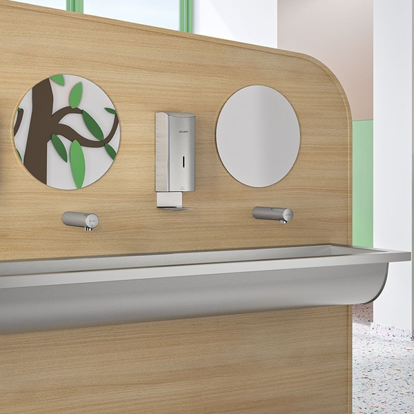 Heller Waschraum mit Edelstahl-Waschrinnbecken, elektronischen Armaturen, Seifenspender und runden Dekorelementen auf einer Holzwand.