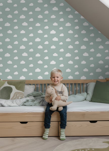 Helles Kinderzimmer mit Vliestapete im Wolkenmuster, Holzbett mit Schubladen und einem Kind, das mit einem Stofftier auf dem Bett sitzt.