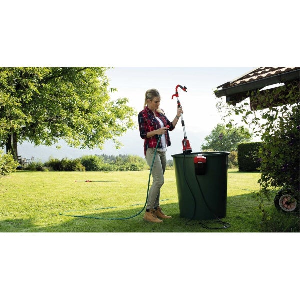 Eine Frau bedient eine batteriebetriebene Einhell Regenfasspumpe an einer Regentonne in einem sonnigen Garten zur Bewässerung mit Regenwasser.