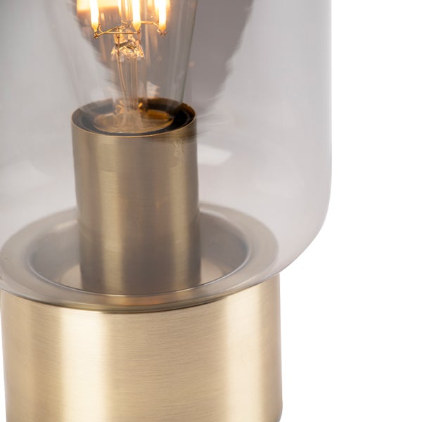 Detail einer Tischleuchte mit Sockel aus gebürstetem Messing, Schirm aus klarem Glas und leuchtendem Leuchtmittel.