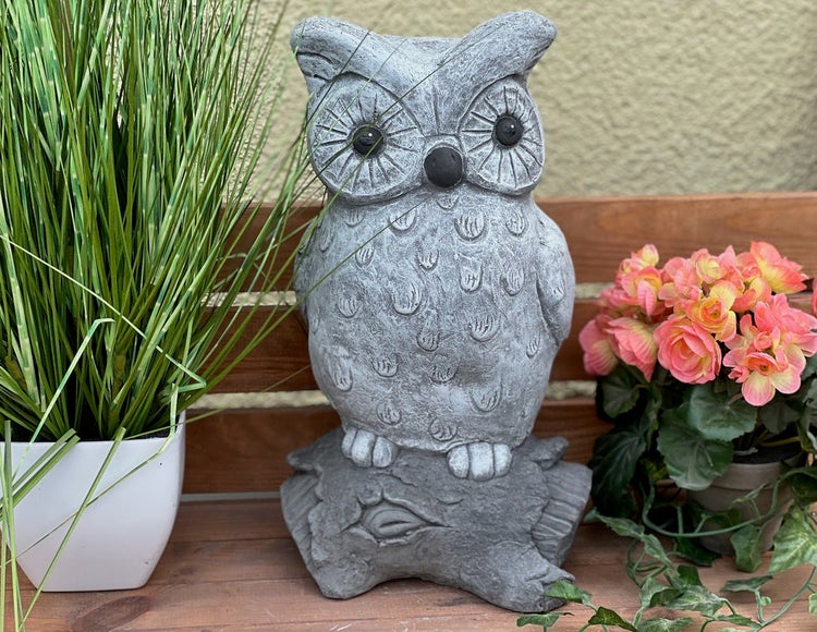 Gartenfigur in Form einer Eule in Steinoptik, sitzend auf einem Baumstamm, dekorativ platziert zwischen Topfpflanzen auf einer Holzbank.
