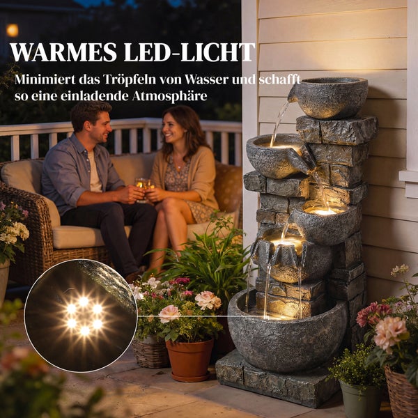 Hell gestalteter Gartenbrunnen in Natursteinoptik mit kaskadenförmigen Schalen und Beleuchtung zwischen Blumen im Garten.