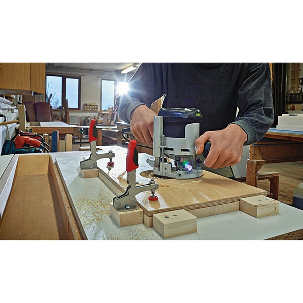 Eine Person verwendet eine Oberfräse mit Zwingen an einem Holzstück in einer Werkstatt.