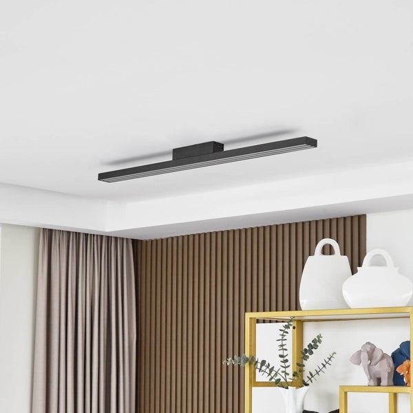 Moderne, längliche LED-Deckenleuchte in Schwarz an einer weißen Decke über einer Wand mit Holzlamellen und einem goldenen Regal.