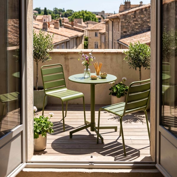 Moderner Balkon mit rundem Metalltisch, zwei Metallstühlen und Holzdielen. Helle Szene mit Topfpflanzen und Blick auf Dächer der Stadt.