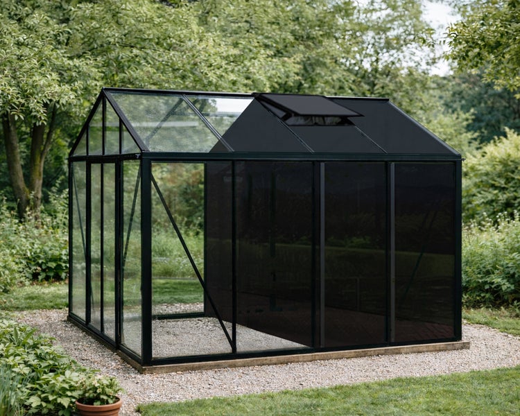 Gewächshaus mit schwarzem Aluminiumrahmen, Glaswänden und schwarzer Sonnenschutzfolie auf einer Kiesfläche im Garten.