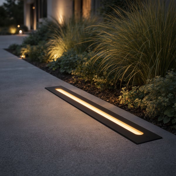 Eine lineare LED-Bodeneinbauleuchte beleuchtet einen Gartenweg aus Beton neben einer Bepflanzung mit Gräsern.