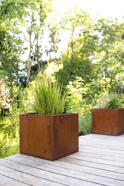 Quadratische Pflanzkübel aus Cortenstahl auf einer Holzterrasse in einem sonnigen Garten.