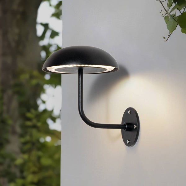 Schwarze Außenwandleuchte in Pilzform aus Metall, an einer weißen Wand montiert, strahlt warmes Licht nach unten ab.