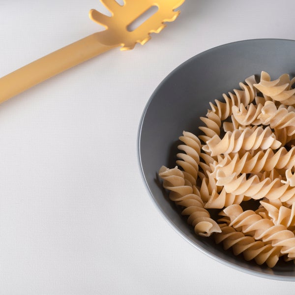 Graue Schüssel mit Fusilli-Nudeln und ein gelber Pastalöffel auf einem weißen Tischtuch.