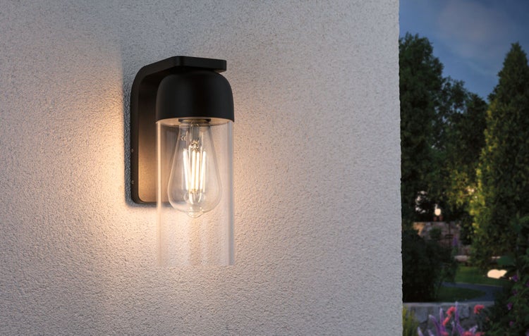 Moderne Außenwandleuchte mit schwarzer Halterung, klarem Glaszylinder und LED-Leuchtmittel an einer Hausfassade mit Garten im Hintergrund.