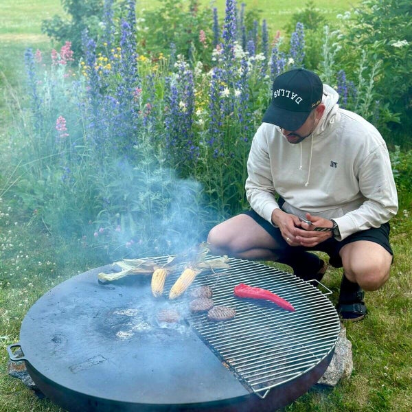 Ein Mann grillt Mais, Frikadellen und Paprika auf einer runden Feuerschale mit Grillrost und Platte in einem blühenden Garten.