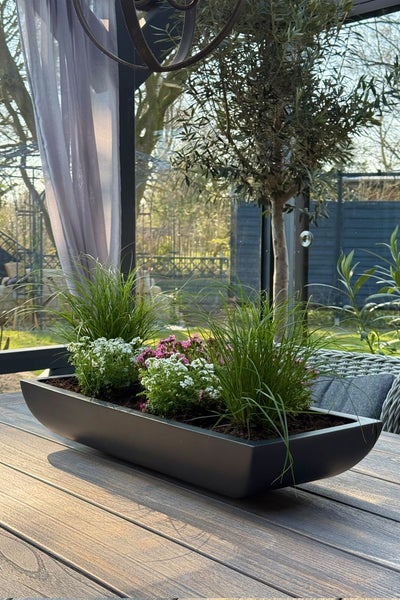Moderner, bootsförmiger Pflanzkübel mit Ziergräsern sowie weißen und rosa Blumen auf einem Holztisch im Außenbereich.