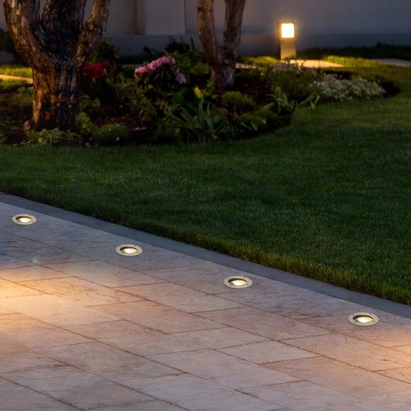 Bodeneinbaustrahler auf einer gepflasterten Terrasse im Garten bei Nacht mit Rasen und beleuchteten Bäumen.